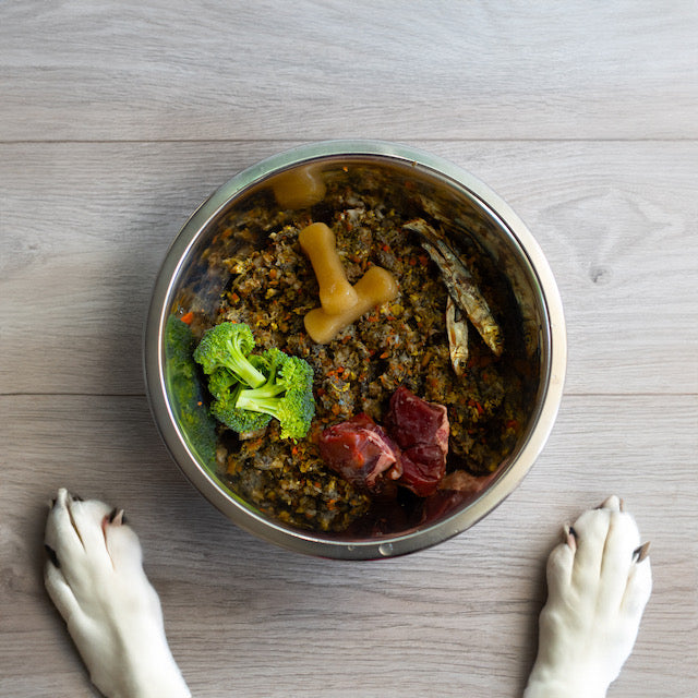 Overhead photo of silver dog bowl filled with fresh cuts of meat, brocolli, sprats and two small frozen bone broth Happy Bones. Bowl is placed on a wooden floor with two white dog paws poking in from the bottom of the shot.