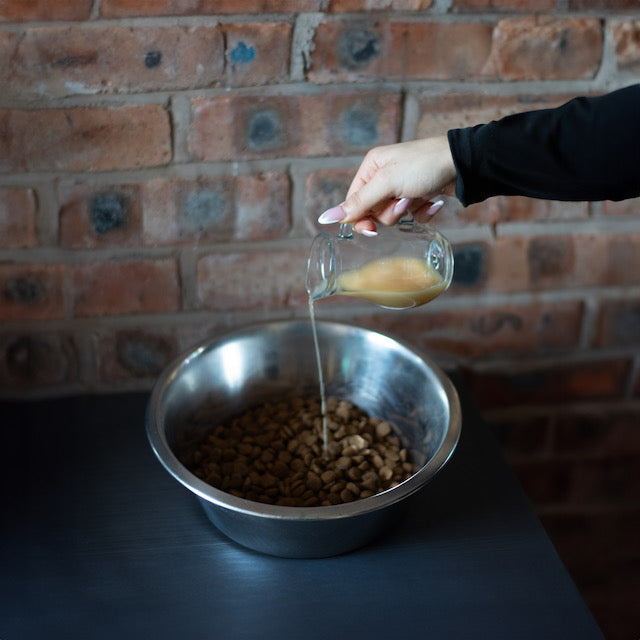 Femal hand pouring bone broth from a clear glass jug over dry dog ibble n a silver dog bowl. The bowl is sat on a dark work surface against an exposed brick wall.